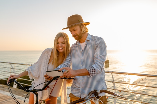 Giovane attraente sorridente felice uomo e donna che viaggiano in bicicletta utilizzando smartphone, coppia romantica in riva al mare sul tramonto, vestito stile boho hipster, amici che si divertono insieme