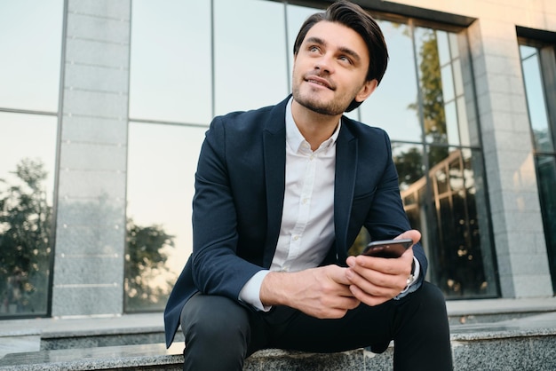 Giovane attraente uomo bruna barbuto sorridente in camicia bianca e abito classico che guarda sognante da parte tenendo il cellulare in mano con edificio in vetro sullo sfondo