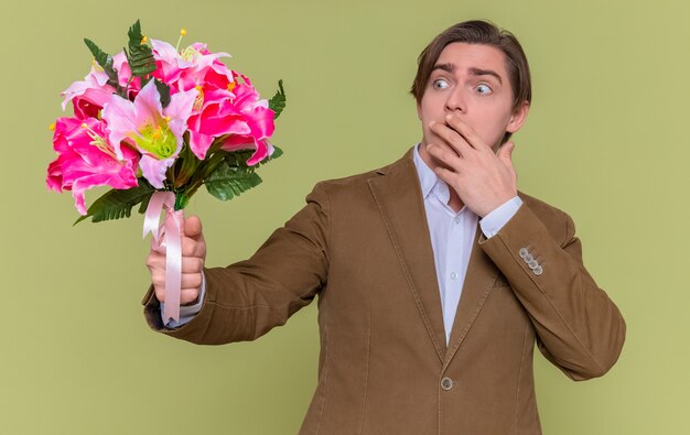 giovane azienda bouquet di fiori guardandolo sorpreso che copre la bocca con la mano