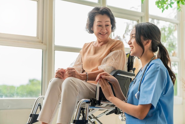 Giovane badante asiatica che si prende cura del suo paziente anziano all'asilo nido Paziente handicappato su una sedia a rotelle in ospedale che parla con un'infermiera amichevole e che guarda un'infermiera allegra che si sposta sul paziente anziano