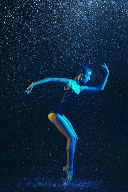 Giovane ballerina femminile che si esibisce sotto gocce d'acqua e spray. Modello caucasico che balla in luci al neon. Donna attraente. Concetto di balletto e coreografia contemporanea. Foto d'arte creativa.