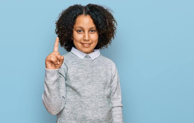 Giovane bambina con i capelli afro che indossa abiti casual mostrando e puntando verso l'alto con il dito numero uno mentre sorride fiducioso e felice