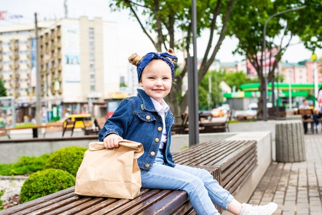 Giovane bambina con la borsa degli alimenti a rapida preparazione vicino al caffè che si siede sulla panchina