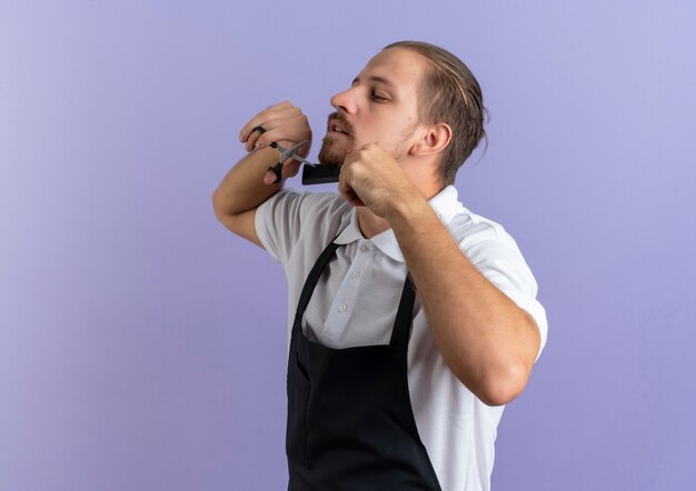 Giovane barbiere bello che indossa la pettinatura uniforme e il taglio della propria barba isolato su sfondo viola con spazio di copia