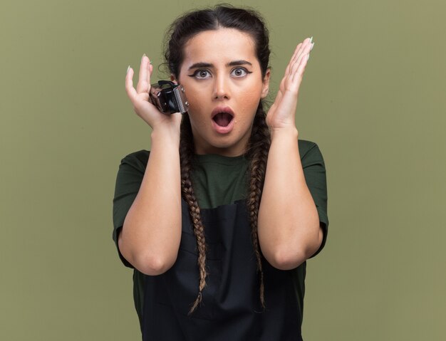 Giovane barbiere femminile sorpreso in tagliatori di capelli della tenuta uniforme che tengono le mani intorno al fronte isolato sulla parete verde oliva