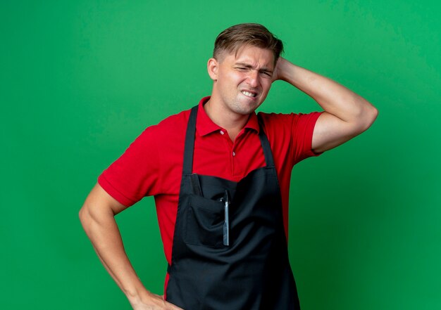 Giovane barbiere maschio biondo ansioso in uniforme morde le labbra mettendo la mano dietro la testa isolata su uno spazio verde con spazio di copia