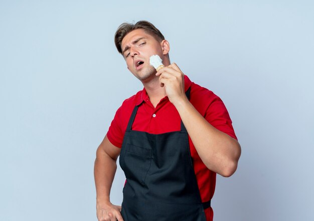 Giovane barbiere maschio biondo fiducioso in schiuma da barba sbavature uniforme sul viso con pennello da barba isolato su spazio bianco con lo spazio della copia