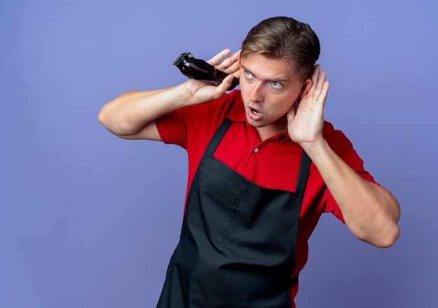 Giovane barbiere maschio biondo gioioso in uniforme mette le mani dietro le orecchie guardando il tagliatore di capelli della holding del lato isolato sullo spazio viola con lo spazio della copia