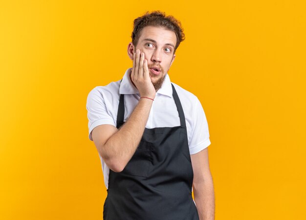 Giovane barbiere maschio dall'aspetto confuso che indossa l'uniforme mettendo la mano sulla guancia isolata sul muro giallo