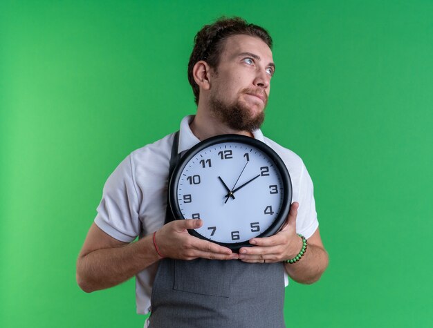 Giovane barbiere maschio dall'aspetto impressionato che indossa un orologio da parete con tenuta in uniforme isolato su parete verde