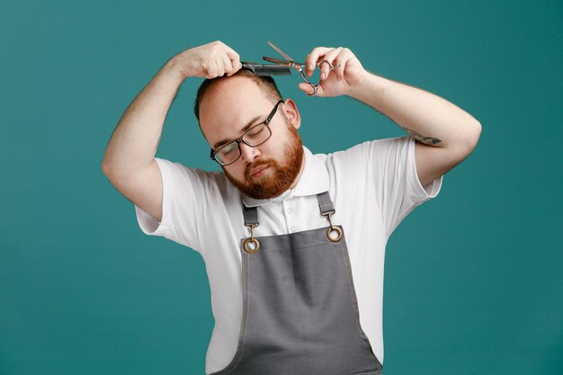 Giovane barbiere serio che indossa uniforme e occhiali con pettine teaser e forbici che si tagliano i capelli con gli occhi chiusi isolati su sfondo blu