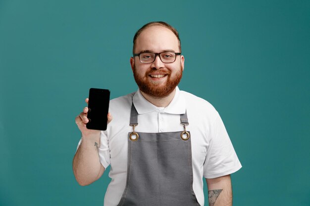 Giovane barbiere sorridente che indossa uniforme e occhiali che mostrano il telefono cellulare mentre guarda la fotocamera isolata su sfondo blu