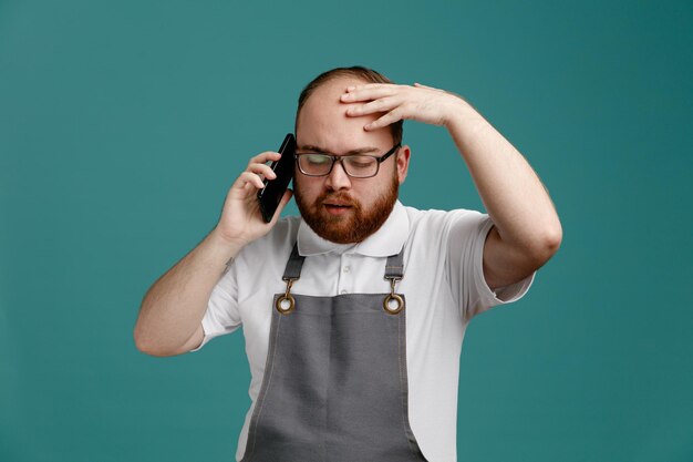 Giovane barbiere stanco che indossa uniforme e occhiali tenendo la mano sulla testa parlando al telefono con gli occhi chiusi isolati su sfondo blu