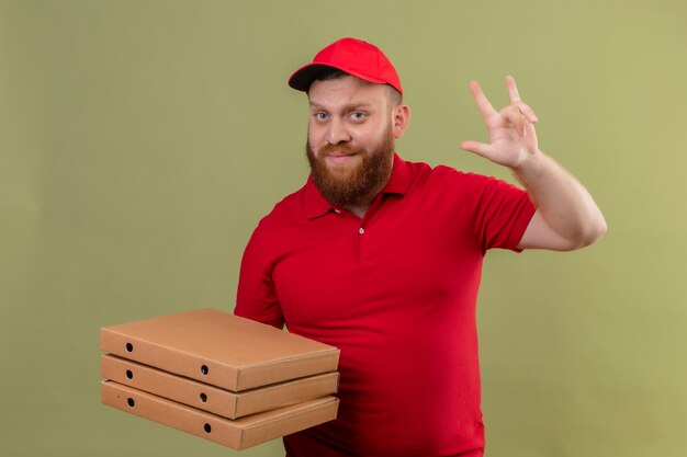 Giovane barbuto fattorino in uniforme rossa e cappuccio che tiene pila di scatole per pizza cercando fiducioso facendo il simbolo del rock