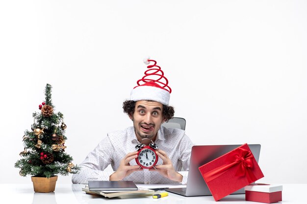 Giovane barbuto uomo d'affari divertente con cappello di Babbo Natale e mostrando orologio guardando qualcosa di sorpresa e seduto in ufficio su sfondo bianco