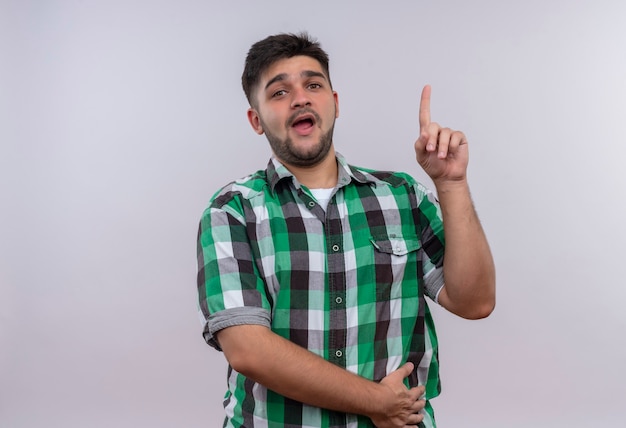 Giovane bel ragazzo che indossa la camicia a scacchi guardando sorpreso rivolto verso l'alto con l'indice in piedi sopra il muro bianco