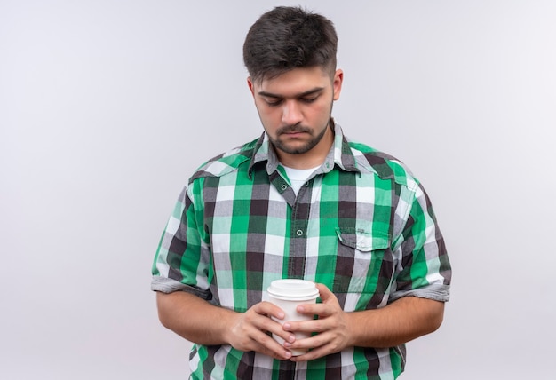 Giovane bel ragazzo che indossa la camicia a scacchi guardando tristemente verso il basso sulla sua tazza di caffè di plastica in piedi sopra il muro bianco