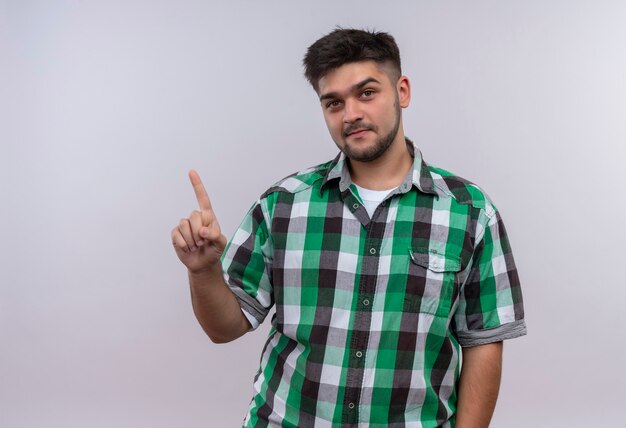 Giovane bel ragazzo che indossa la camicia a scacchi sorridente rivolto verso l'alto con l'indice in piedi sopra il muro bianco