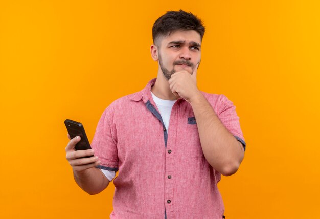 Giovane bel ragazzo che indossa la maglietta polo rosa guardando oltre premurosamente tenendo il telefono in piedi sopra la parete arancione