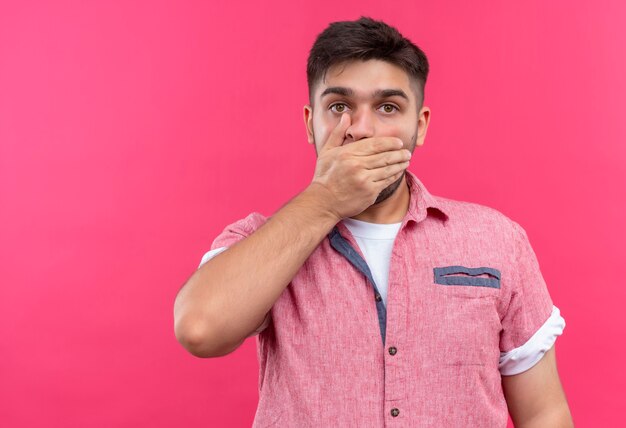 Giovane bel ragazzo che indossa la maglietta polo rosa guardando scioccato in piedi sopra il muro rosa