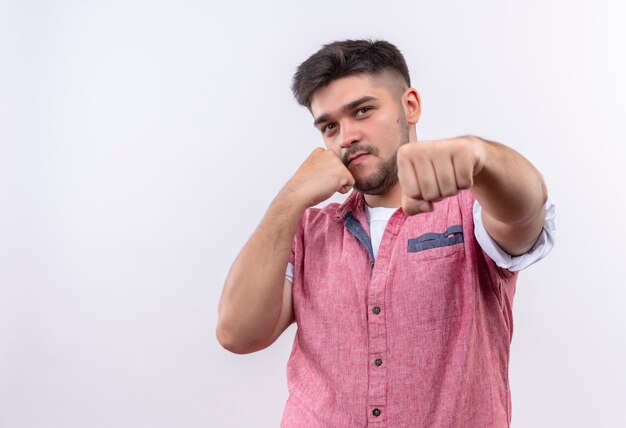 Giovane bel ragazzo che indossa la polo rosa mostra la boxe in piedi sul muro bianco