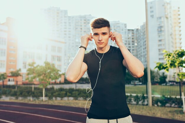 Giovane bel ragazzo con un buon corpo la mattina sullo stadio. Indossa una maglietta nera, mette le cuffie alle orecchie e guarda la telecamera.