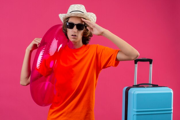 Giovane bel ragazzo in maglietta arancione e cappello estivo indossando occhiali da sole neri tenendo l'anello gonfiabile toccando la testa per errore cercando confuso in piedi con la valigia di viaggio sul backgrou rosa