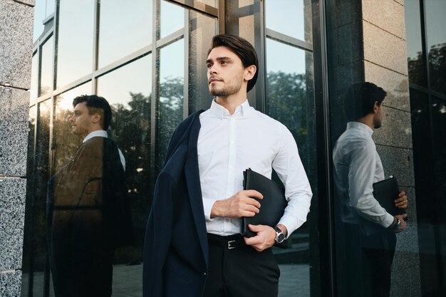 Giovane bell'uomo bruna con la barba in camicia bianca e giacca classica sulla spalla che guarda sognante da parte con la borsa per i documenti in mano e la costruzione di vetro sullo sfondo