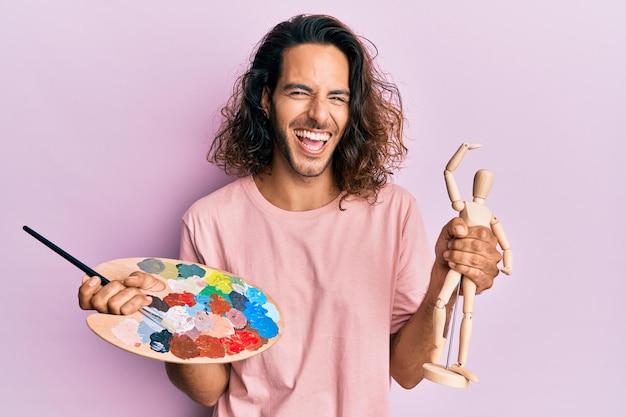 Giovane bell'uomo con i capelli lunghi che tiene la tavolozza del pittore e il manichino d'arte che sorride e ride ad alta voce perché divertente scherzo pazzo.