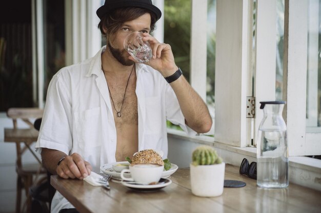 Giovane bell'uomo con una camicia bianca aperta, fare colazione in un bar con un hamburger vegetariano, bere caffè, stile di vita in un'isola tropicale, vita a Bali.