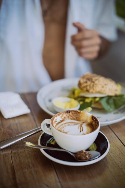 Giovane bell'uomo con una camicia bianca aperta, fare colazione in un bar con un hamburger vegetariano, bere caffè, stile di vita in un'isola tropicale, vita a Bali.