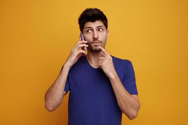 Giovane bell'uomo premuroso che parla al telefono guardando il lato tenendo il dito sul mento su sfondo giallo