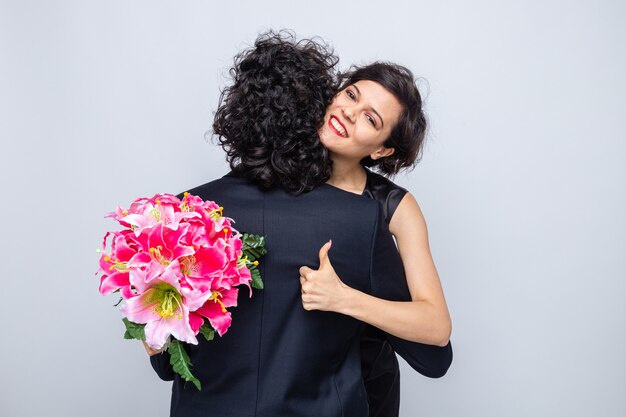 Giovane bella coppia donna felice con un mazzo di fiori che abbraccia il suo ragazzo che mostra i pollici in su sorridendo