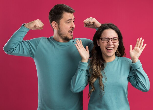 Giovane bella coppia in abiti casual blu uomo e donna felici ed emozionati alzando le braccia e stringendo i pugni felici innamorati insieme in piedi sul muro rosa