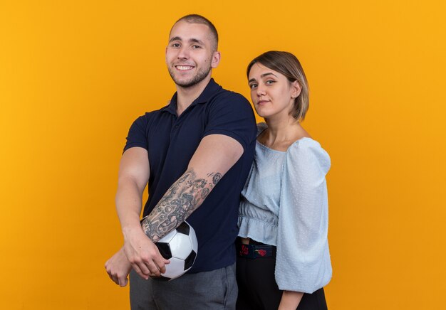 Giovane bella coppia in abiti casual uomo con pallone da calcio e donna che sembra felice e positiva sorridente allegramente in piedi