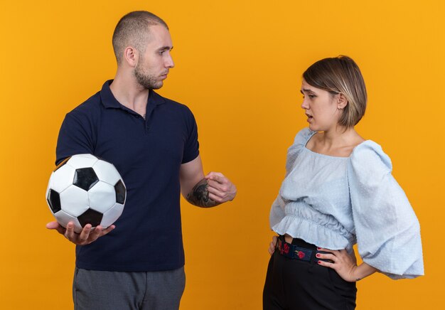 Giovane bella coppia in abiti casual uomo con pallone da calcio guardando la sua ragazza confusa e dispiaciuta in piedi