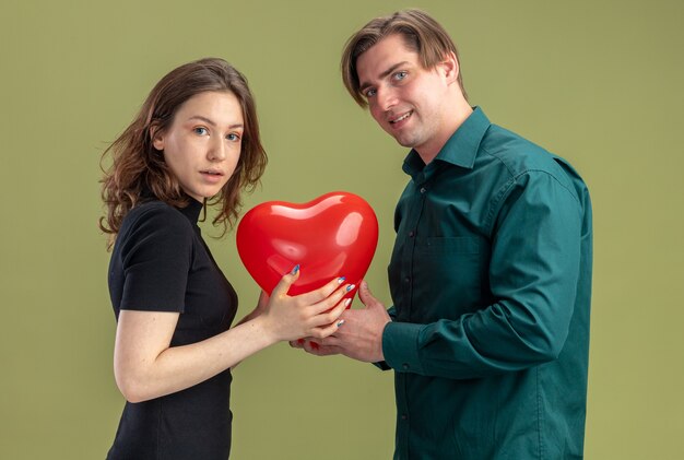 Giovane bella coppia in abiti casual uomo e donna con palloncino a forma di cuore felice innamorato insieme abbracciando per celebrare il giorno di San Valentino in piedi su sfondo verde