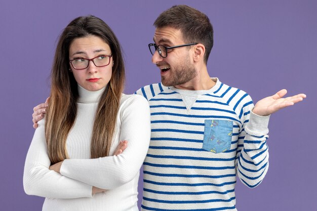 giovane bella coppia in abiti casual uomo sorridente guardando la sua ragazza triste concetto di San Valentino in piedi sul muro viola