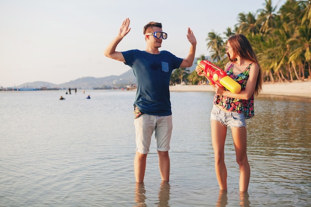 Giovane bella coppia innamorata che gioca sulla spiaggia tropicale, vacanze estive, luna di miele, romanticismo, tramonto, felice, divertirsi, pistola ad acqua, lotta, l'uomo si arrende, positivo, divertente