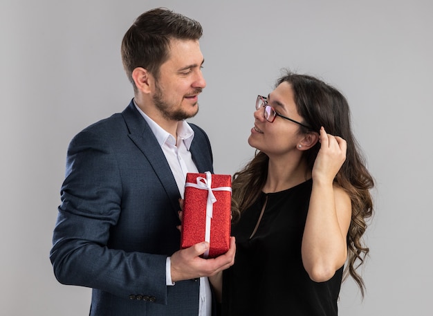 Giovane bella coppia uomo che tiene un regalo per la sua allegra fidanzata felice innamorata che celebra il giorno di San Valentino in piedi sul muro bianco