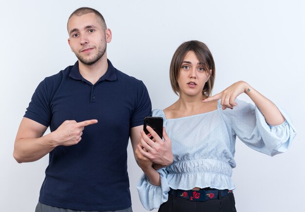 Giovane bella coppia uomo e donna che guarda l'uomo con lo smartphone che punta con il dito indice alla sua ragazza confusa in piedi