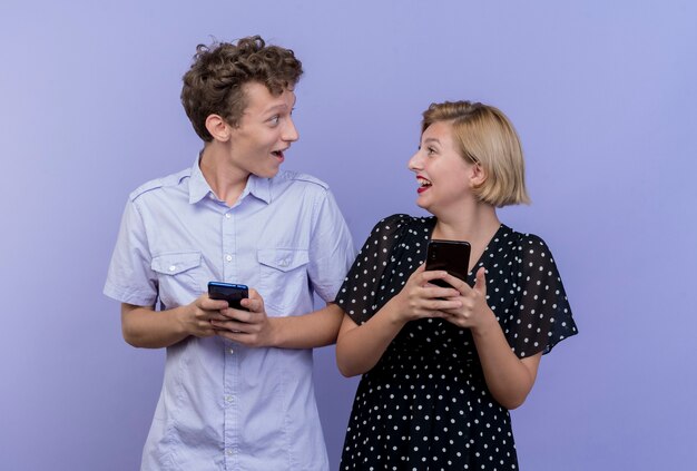 Giovane bella coppia uomo e donna che tengono gli smartphone guardando a vicenda con il sorriso in piedi sopra la parete blu