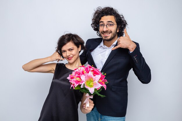 Giovane bella coppia uomo e donna con bouquet di fiori che fa mi chiama gesto sorridendo allegramente felice in amore che celebra la giornata internazionale della donna 8 marzo