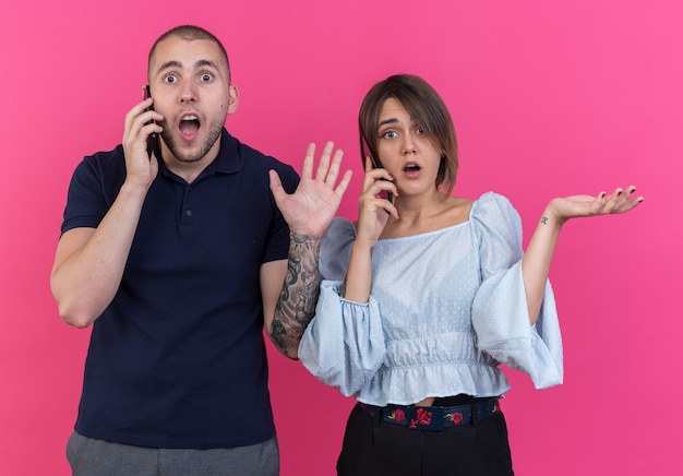 Giovane bella coppia uomo e donna confusa con le braccia alzate mentre parla al cellulare in piedi sul muro rosa