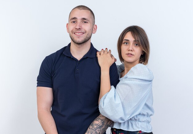 Giovane bella coppia uomo e donna felice e positiva donna sorridente che tiene le mani sulla spalla del suo ragazzo in piedi sul muro bianco