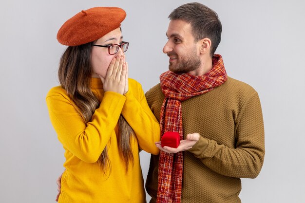 Giovane bella coppia uomo felice che fa proposta con anello di fidanzamento nel riquadro rosso alla sua ragazza stupita in berretto durante il giorno di San Valentino in piedi su sfondo bianco