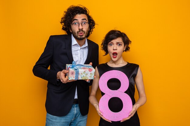 Giovane bella coppia uomo felice con regalo e donna con numero otto guardando la telecamera stupita e sorpresa celebrando la giornata internazionale della donna 8 marzo in piedi su sfondo arancione