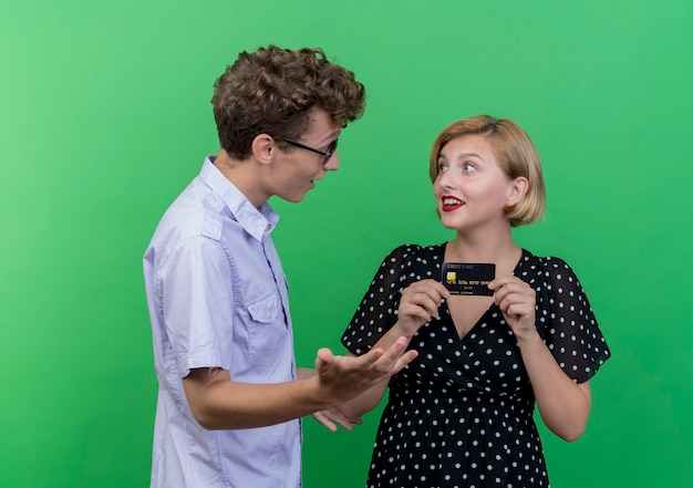 Giovane bella coppia uomo sorpreso guardando la ragazza felice che tiene la carta di credito in piedi sopra la parete verde