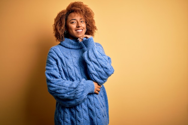 Giovane bella donna afroamericana afroamericana con i capelli ricci che indossa un maglione a collo alto blu che guarda fiducioso alla telecamera con un sorriso con le braccia incrociate e la mano alzata sul mento Pensando positivo