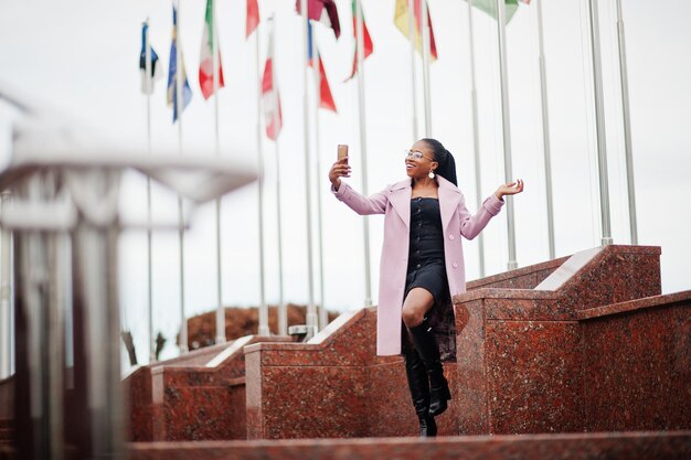 Giovane bella donna afroamericana alla moda in strada che indossa il cappotto dell'abito di moda contro le bandiere di diversi paesi del mondo con il telefono cellulare a portata di mano